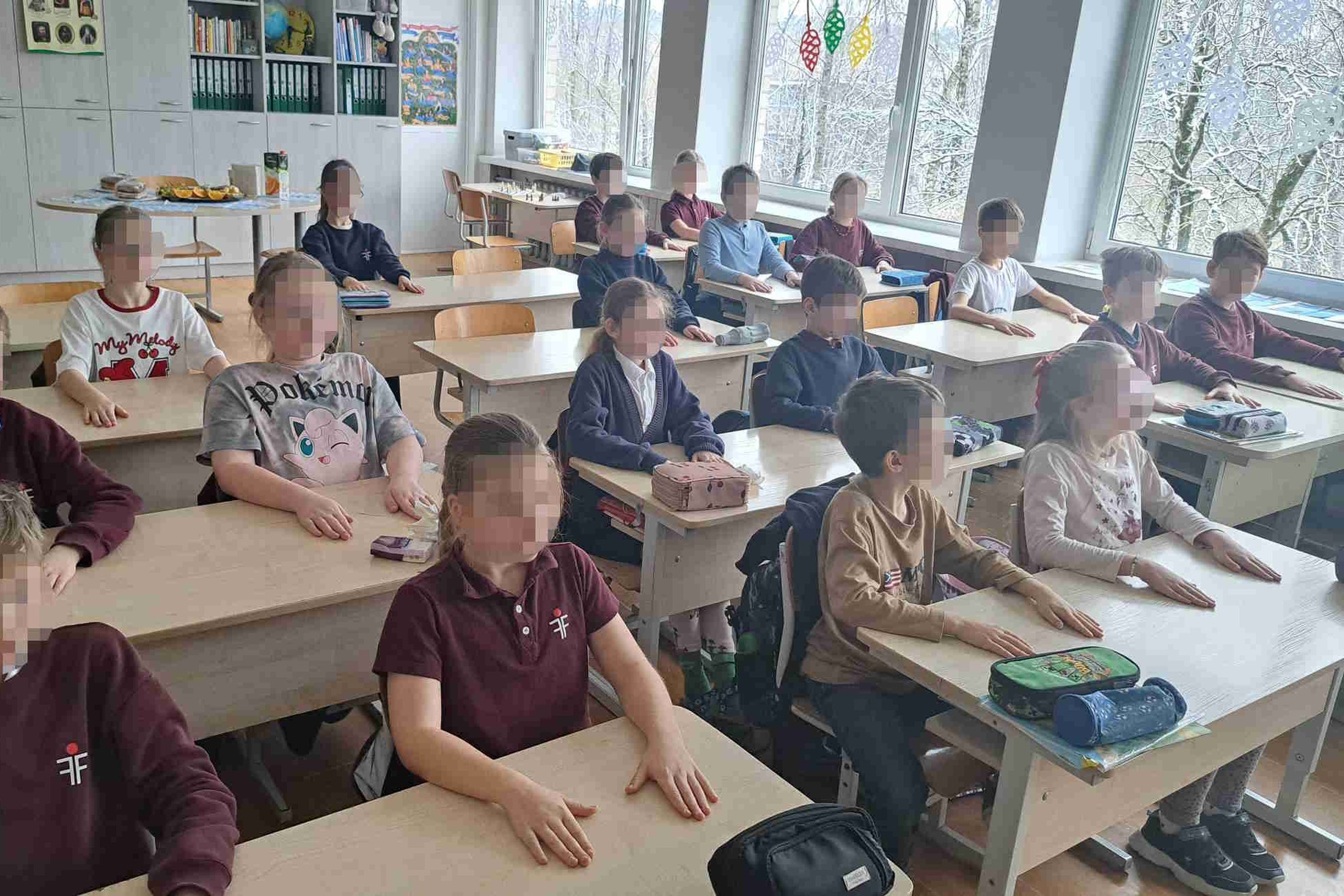 Vaikai sėdi klasėje prie savo suolų tvarkingomis eilėmis, visi nuleidę rankas ant stalų, žvelgia į priekį; fone matyti lentynos su knygomis ir užkandžių stalas, pro langus atsiveria ankstyvojo pavasario peizažas.