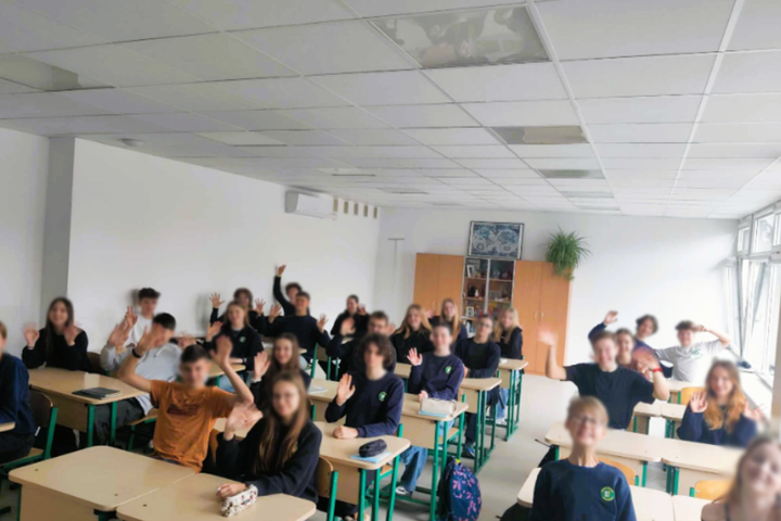 Paveikslėlyje matoma klasė su moksleiviais, sėdinčiais prie stalų ir mojuojančiais į kamerą. Vaikai atrodo linksmi ir įsitraukę, dauguma vilki vienodą tamsų aprangą – galimai mokyklinę uniformą. Klasėje šviesu, pro didelius langus sklinda natūrali šviesa,