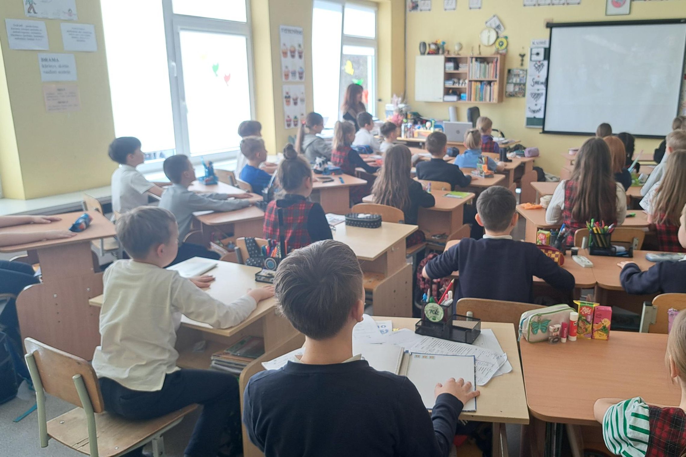 Pradinukai sėdi savo suoluose šviesioje klasėje, žiūrėdami į projekcinį ekraną, kuriame rodomas vaizdas; kai kurie vaikai vilki languotus uniforminius sarafanus, ant stalų – sąsiuviniai, penalai ir piešimo priemonės, o fone – lentynos su knygomis ir mokyt