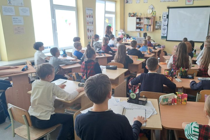 Pradinukai sėdi savo suoluose šviesioje klasėje, žiūrėdami į projekcinį ekraną, kuriame rodomas vaizdas; kai kurie vaikai vilki languotus uniforminius sarafanus, ant stalų – sąsiuviniai, penalai ir piešimo priemonės, o fone – lentynos su knygomis ir mokyt