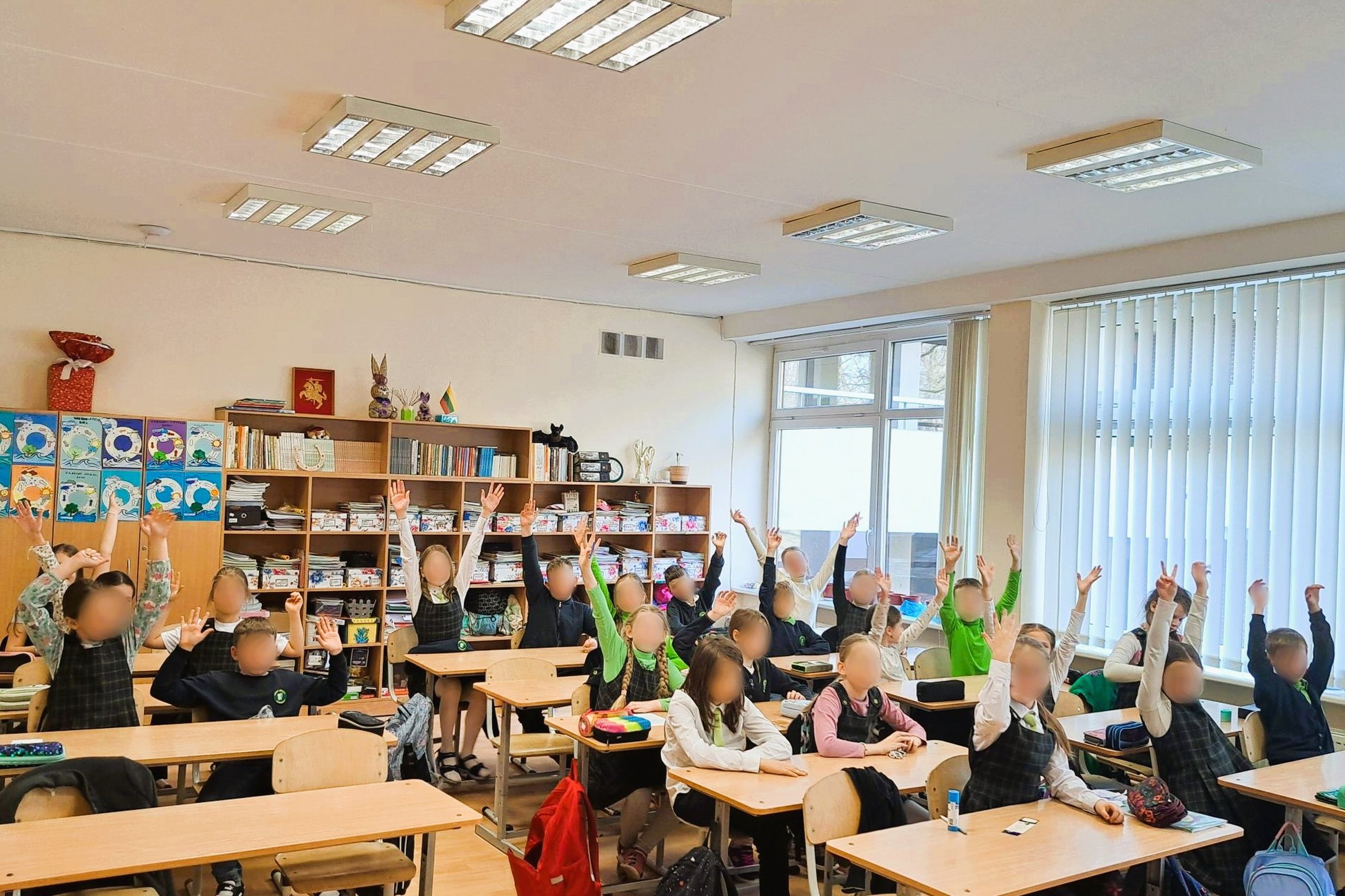 Pradinės mokyklos klasėje sėdi vaikai, dauguma jų pakėlę rankas. Jie vilki tamsius languotus uniforminius sarafanus arba megztinius su žaliu akcentu. Klasė šviesi, tvarkinga, su knygų lentynomis ir mokinių darbais ant sienų. Vaikų rankų pakėlimas rodo akt