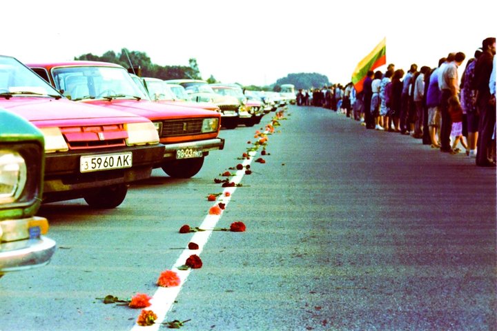 Istorinė nuotrauka iš Baltijos kelio – žmonės susikibę rankomis stovi kelkraščiu, sudarydami gyvą grandinę. Kitoje kelio pusėje stovi sovietiniai automobiliai, o per kelio vidurį išrikiuotos gėlės. Matomas trispalvės – Lietuvos vėliavos – fragmentas. Vaiz
