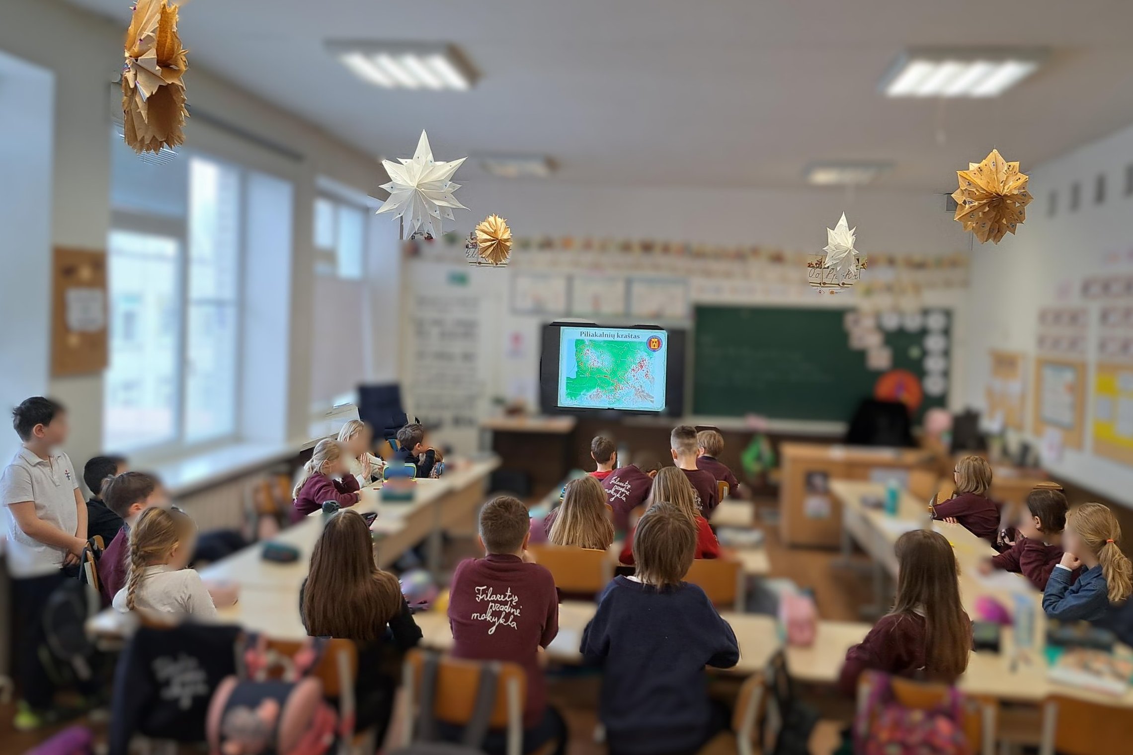 Filaretų pradinės mokyklos mokiniai klasėje žiūri į ekraną, kuriame rodoma pilkapių ir piliakalnių sklaidos schema Europoje; klasės aplinka papuošta žvaigždės formos šventiniais dekorais, vaikai dėvi mokyklinius džemperius su mokyklos logotipu.