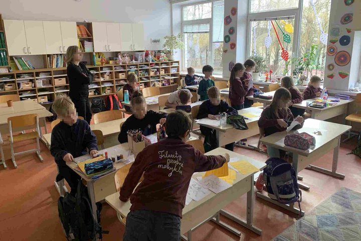 Vaikai klasėje dirba prie savo stalų, piešdami ir kuriantys, o mokytoja stebi ir padeda. Klasėje matomi mokinių piešiniai ir spalvotieji rašikliai. Ant langų kabantys dekoratyviniai papuošalai ir natūralus apšvietimas suteikia jaukią atmosferą.