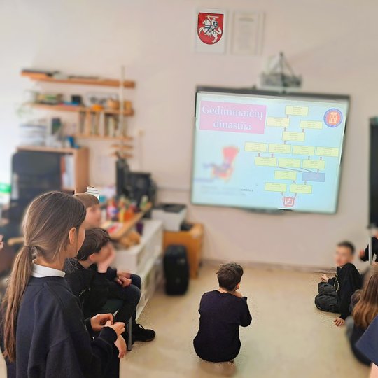 Klasėje vaikai susėdę ant grindų ir stovėdami stebi ant interaktyvios lentos rodomą Gediminaičių dinastijos genealoginį medį. Skaidrėje parodyti Lietuvos valdovų – nuo Gedimino iki Žygimanto Augusto – vardai ir jų tarpusavio giminystės ryšiai. Ant sienos 