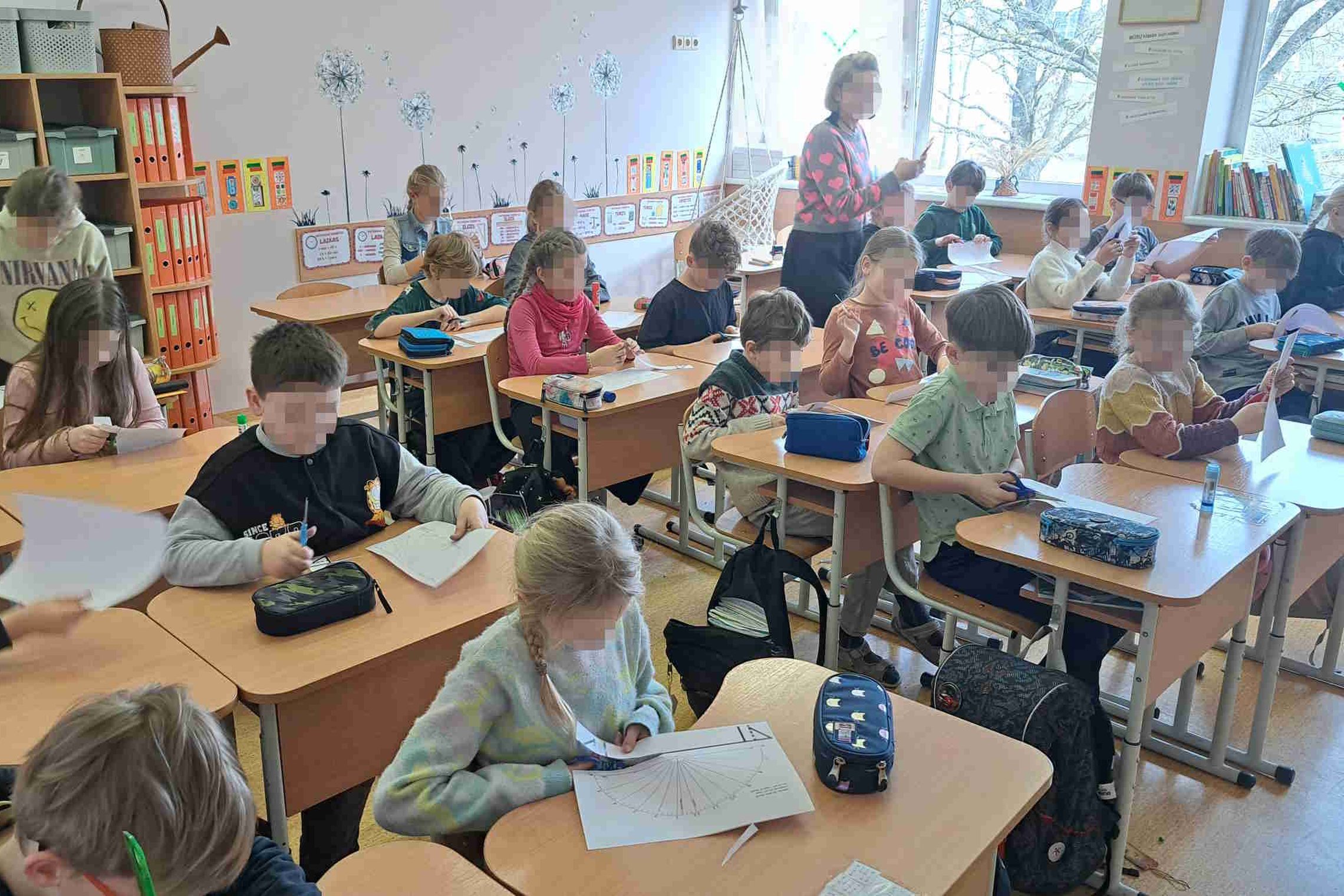 Pradinukai sėdi klasėje prie savo suolų, susikaupę karpo ir pildo laikrodžio formos užduočių lapus. Ant suolų guli penalai, klijai, žirklutės ir sąsiuviniai. Klasės aplinka jauki, su spalvingomis lentynomis ir langais, pro kuriuos matosi medžiai.