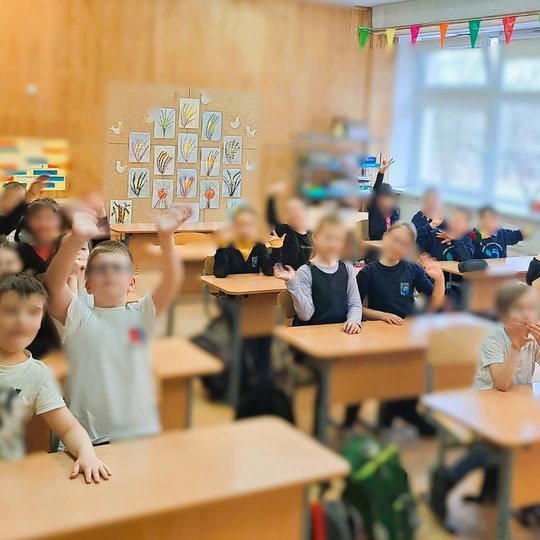 Pradinės klasės mokiniai mojuoja klasėje, fone – vaikų piešinių paroda su augalų motyvais ir spalvinga vėliavėlių girlianda.