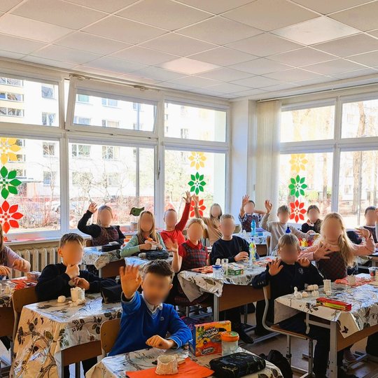 Mokiniai, dirbantys molio edukacijoje klasėje, keliantys rankas ir rodantys savo darbelius. Ant stalo yra įvairių meno reikmenų. Langai dekoruoti spalvotais gėlėmis.