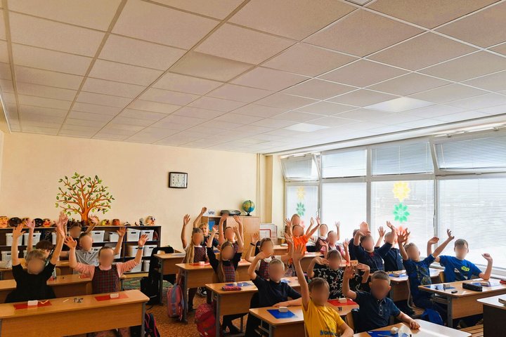  Antrokų klasė, mokiniai pakėlę rankas, atsisėdę prie stalų klasėje, ant sienos matosi laikrodis ir dekoratyvinis medis.
