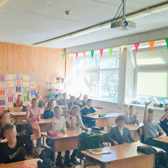 Pradinukai klasėje džiugiai kelia rankas, sėdėdami prie savo suolų šviesioje, spalvingoje mokymosi aplinkoje.
