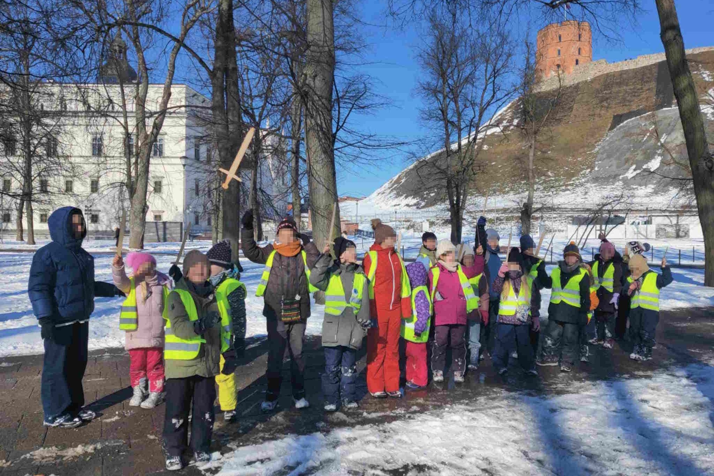 Nuotraukoje matoma grupė vaikų, dėvinčių ryškias saugos liemenes, stovinčių snieguotame parke su Gedimino pilies bokštu fonu Vilniuje. Kai kurie vaikai laiko rankose kalavijus, kas suteikia nuotraukai istorinio konteksto žaismingumo. Ši išvyka atrodo kaip