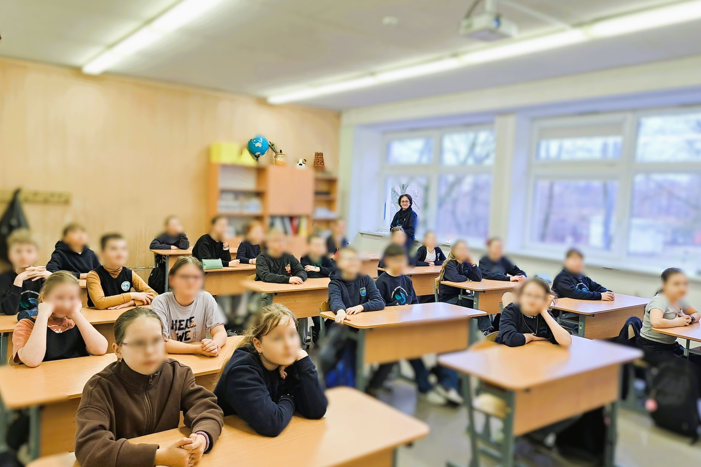Mokytoja ir mokiniai klasėje, sėdintys prie suolų ir dėmesingai klausantys pamokos, pro langus matyti šalto pavasario peizažas.