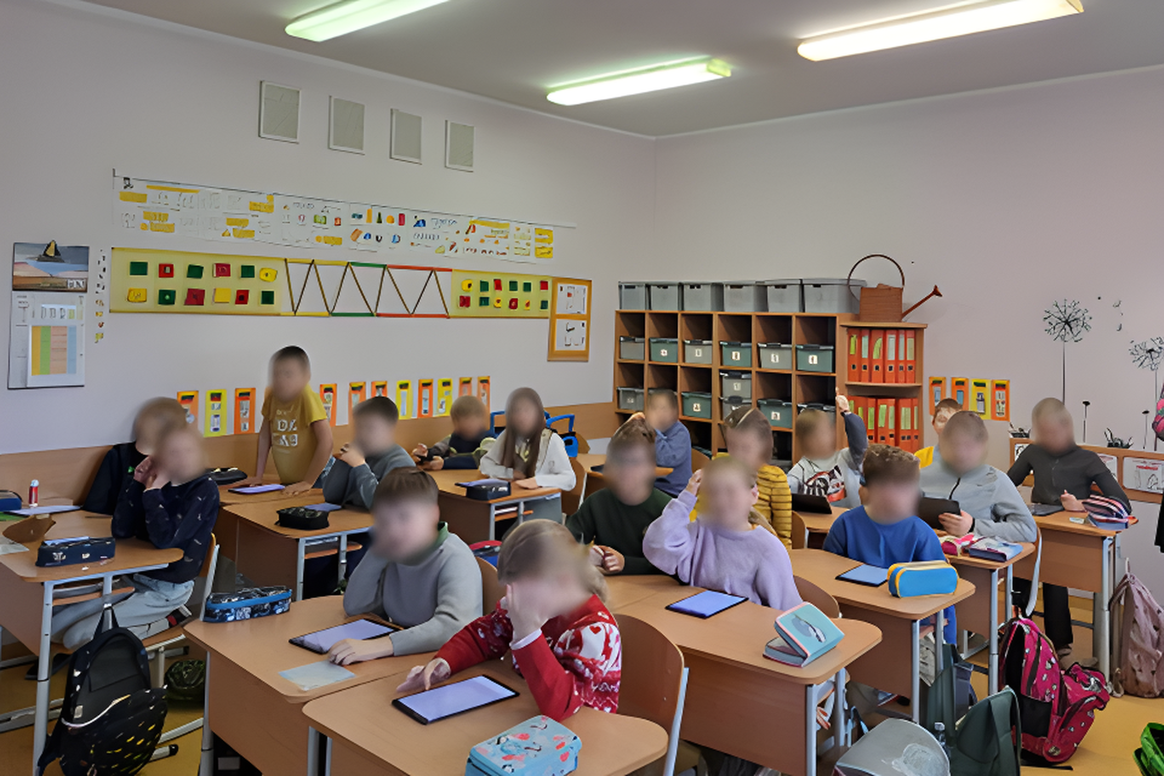 Šioje nuotraukoje vaizduojama klasės aplinka su mokinių grupe, kuri atrodo dalyvaujantys pamokoje. Vaikai sėdi prie savo suoliukų, kuriuose matyti užrašų knygelės ir kiti mokymosi įrankiai. Klasė yra šviesi ir gerai organizuota, su aiškiai išdėstytais mok