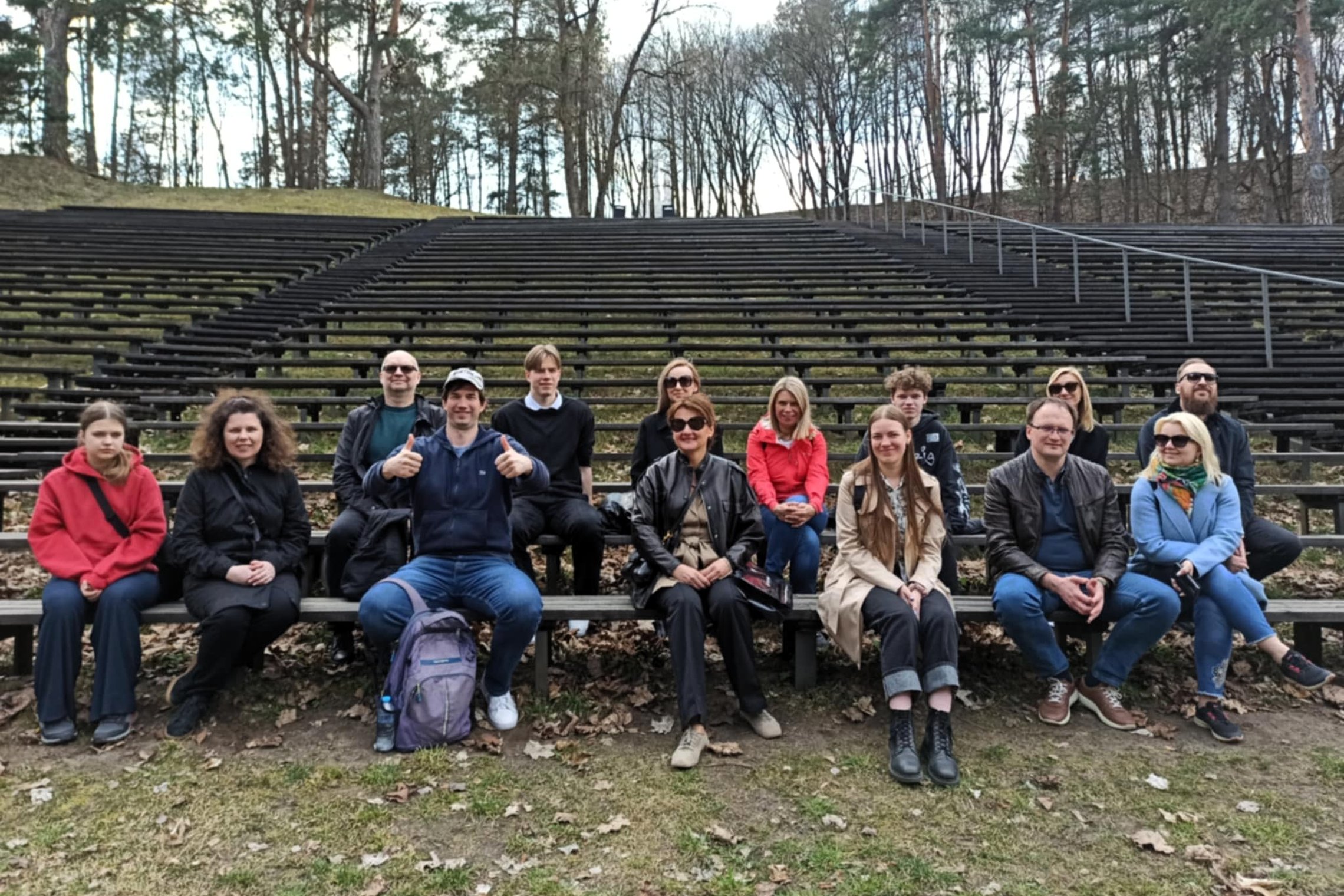 Nuotraukoje matoma grupė žmonių, sėdinčių pirmoje eilėje medinių suolų amfiteatre po atviru dangumi, apsuptame miško. Žmonės šypsosi, dalis pozuoja su pakeltais nykščiais, vyrauja laisvalaikio apranga. Fone matyti tušti suolai, kylantys į kalną, o aplink