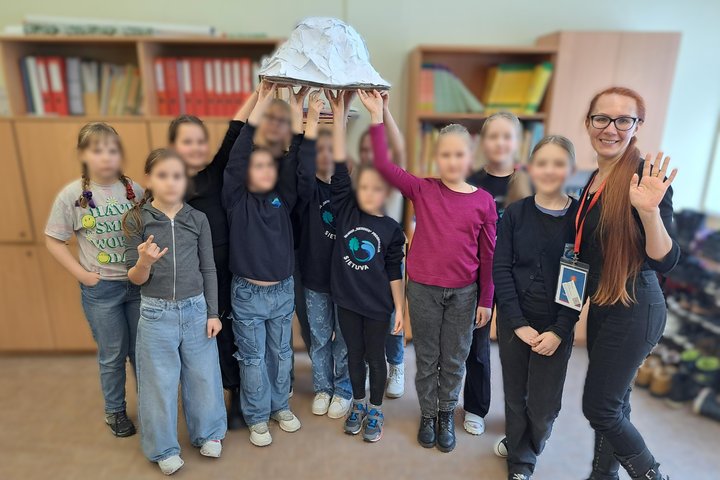 Grupė vaikų klasėje kartu su edukatore pozuoja nuotraukai, laikydami virš galvų bendrą kūrybinį darbą – baltą kupolo formos maketą; fone matyti knygų ir segtuvų lentynos, o edukatorė stovi dešinėje pusėje, mojuodama ir šypsodamasi.