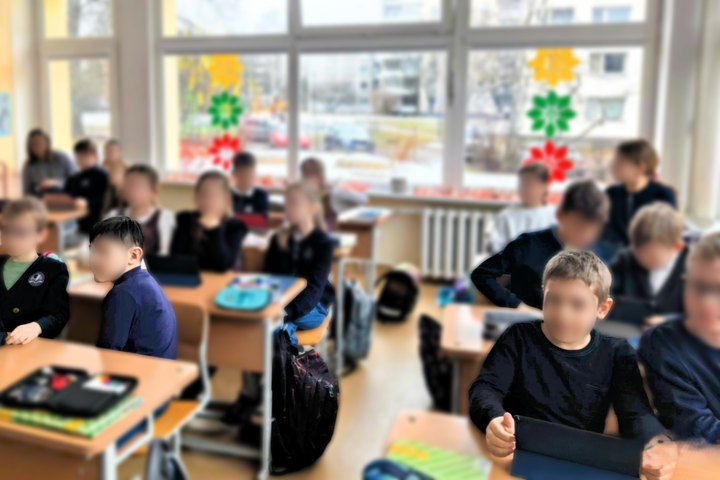 Mokyklos klasėje mokiniai, vilkintys mokyklines uniformas, sėdi prie stalų, kai kurie naudojasi planšetėmis. Pro didelius langus, puoštus spalvotais popieriniais paukščiais, matyti miesto peizažas.