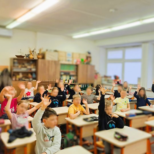 Pradinukai klasėje pakėlę rankas, aplinkoje – medinės spintelės, lentynos su edukacinėmis priemonėmis ir dideli langai.