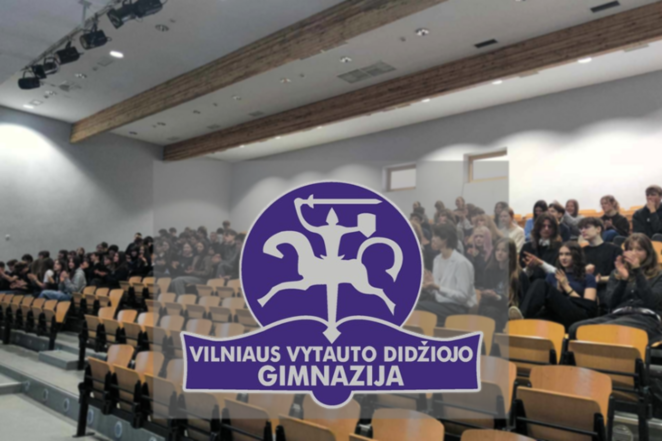 Vilniaus Vytauto Didžiojo gimnazijos aktų salėje sėdi mokiniai, žiūrintys į priekį ir plojantys. Priekyje matomas gimnazijos logotipas su raiteliu ir užrašu „Vilniaus Vytauto Didžiojo gimnazija“.