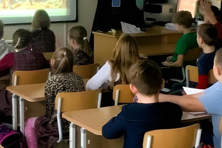 Vaikai klasėje žiūri edukacinį vaizdo įrašą apie senovės Lietuvą, dėmesingai stebi projekciją ir dalyvauja pamokoje.