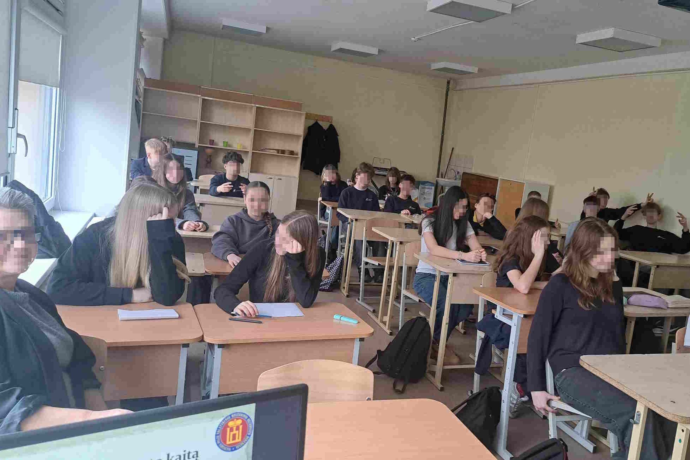 Nuotraukoje matoma klasė paauglių, kurie dalyvauja mokymosi veikloje. Klasės patalpa yra aprūpinta suoliukais ir staliniais su knygomis ir rašymo priemonėmis ant jų. Kai kurie mokiniai dėmesingai žiūri į priekį, kiti šnekučiuojasi su draugais ar tiesiog a