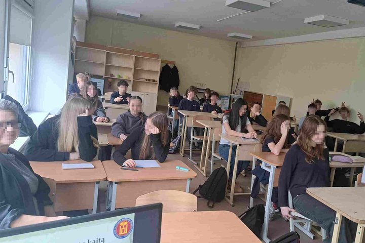 Nuotraukoje matoma klasė paauglių, kurie dalyvauja mokymosi veikloje. Klasės patalpa yra aprūpinta suoliukais ir staliniais su knygomis ir rašymo priemonėmis ant jų. Kai kurie mokiniai dėmesingai žiūri į priekį, kiti šnekučiuojasi su draugais ar tiesiog a