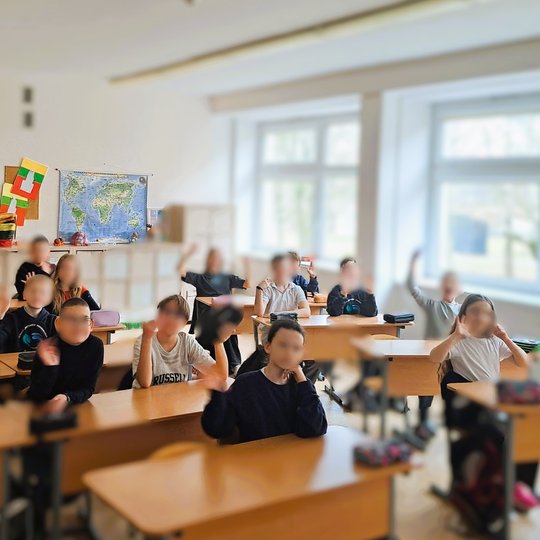 Pradinės mokyklos klasė su mokiniais, sėdinčiais prie suolų ir mojuojančiais, fone kabantys pasaulio ir Lietuvos žemėlapiai bei mokinių rankdarbiai.