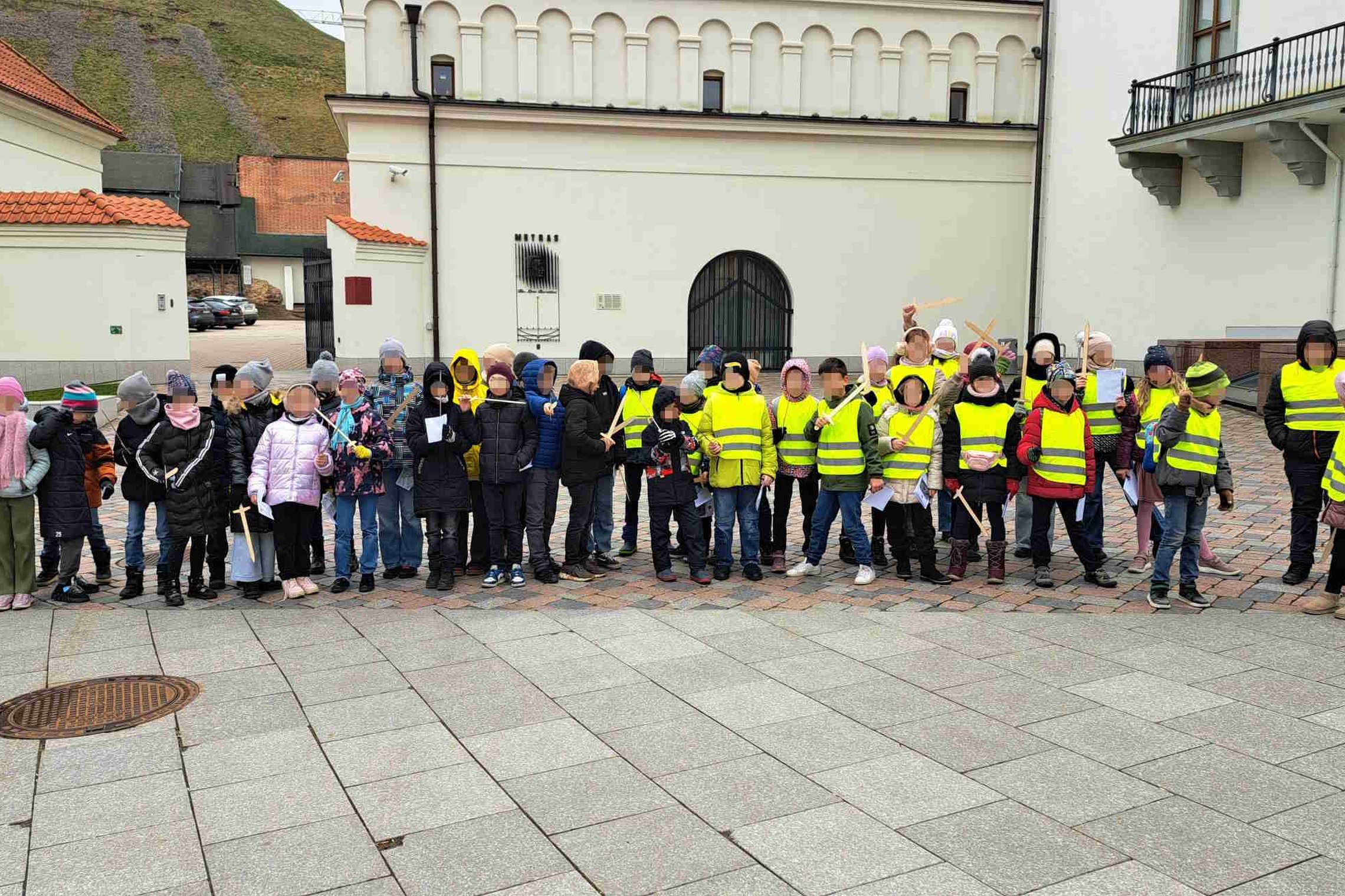 Vaikai su ryškiaspalvėmis liemenėmis stovi prie Valdovų rūmų Vilniaus pilių teritorijoje, laikydami medinius kalavijus ir dalyvaudami edukacinėje veikloje.