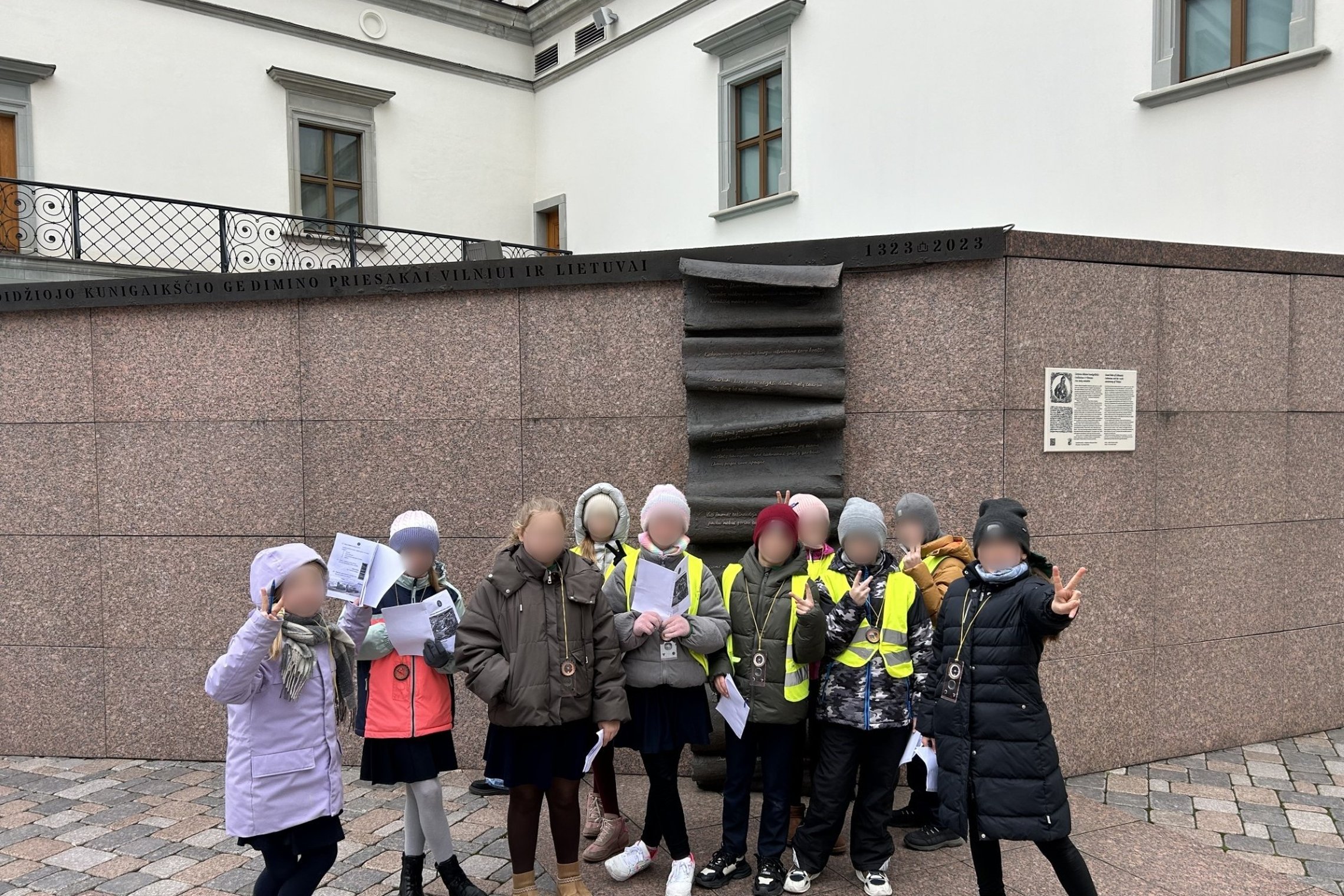 Prie Gedimino tikrojo bokšto sienų, Valdovų rūmų kieme, Vilniaus pilių teritorijoje stovi grupė vaikų su šviesą atspindinčiomis liemenėmis ir edukaciniais ženkliukais ant kaklo. Jie rankose laiko užduočių lapus ir entuziastingai pozuoja, dalis jų rodo per