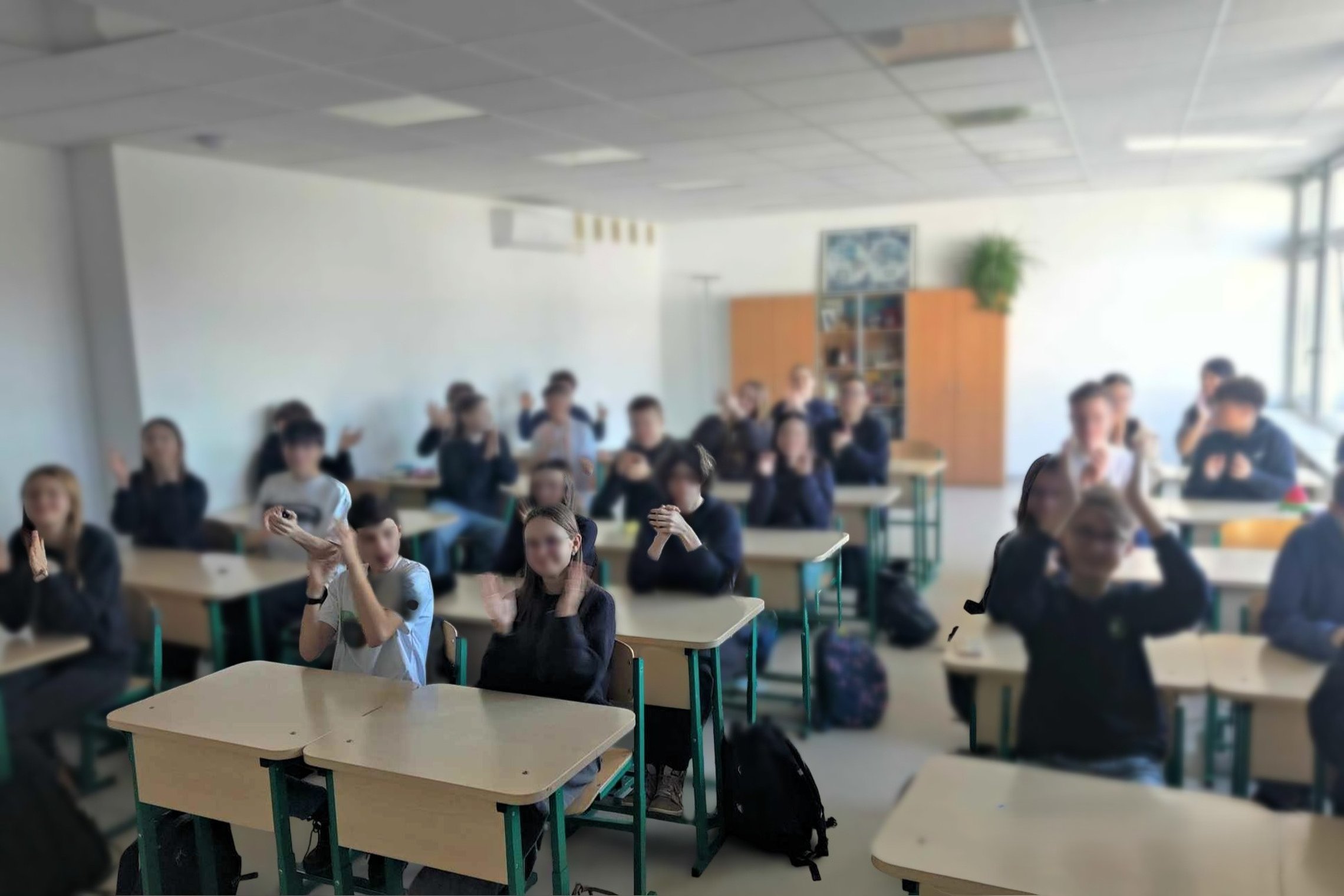 Vidurinės mokyklos mokiniai sėdi klasėje prie savo suolų ir ploja, dalyvaudami pamokoje ar renginyje. Klasės interjeras šviesus, su spintelėmis ir dideliais langais dešinėje pusėje.