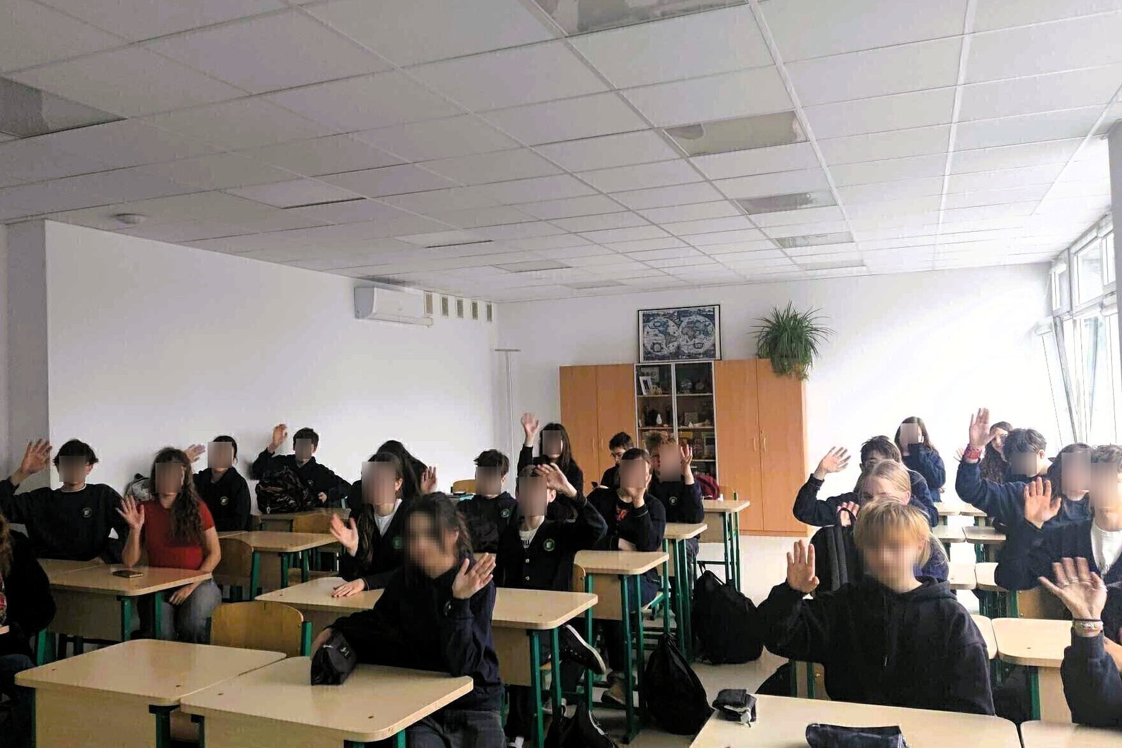 Klasėje prie stalų sėdi mokiniai, iškėlę rankas. Jie vilki tamsiomis uniformomis, klasės gale matyti lentynos ir augalas. Pro didelius langus sklinda natūrali šviesa.
