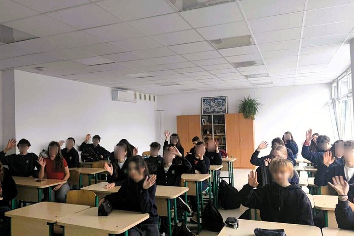 Klasėje prie stalų sėdi mokiniai, iškėlę rankas. Jie vilki tamsiomis uniformomis, klasės gale matyti lentynos ir augalas. Pro didelius langus sklinda natūrali šviesa.