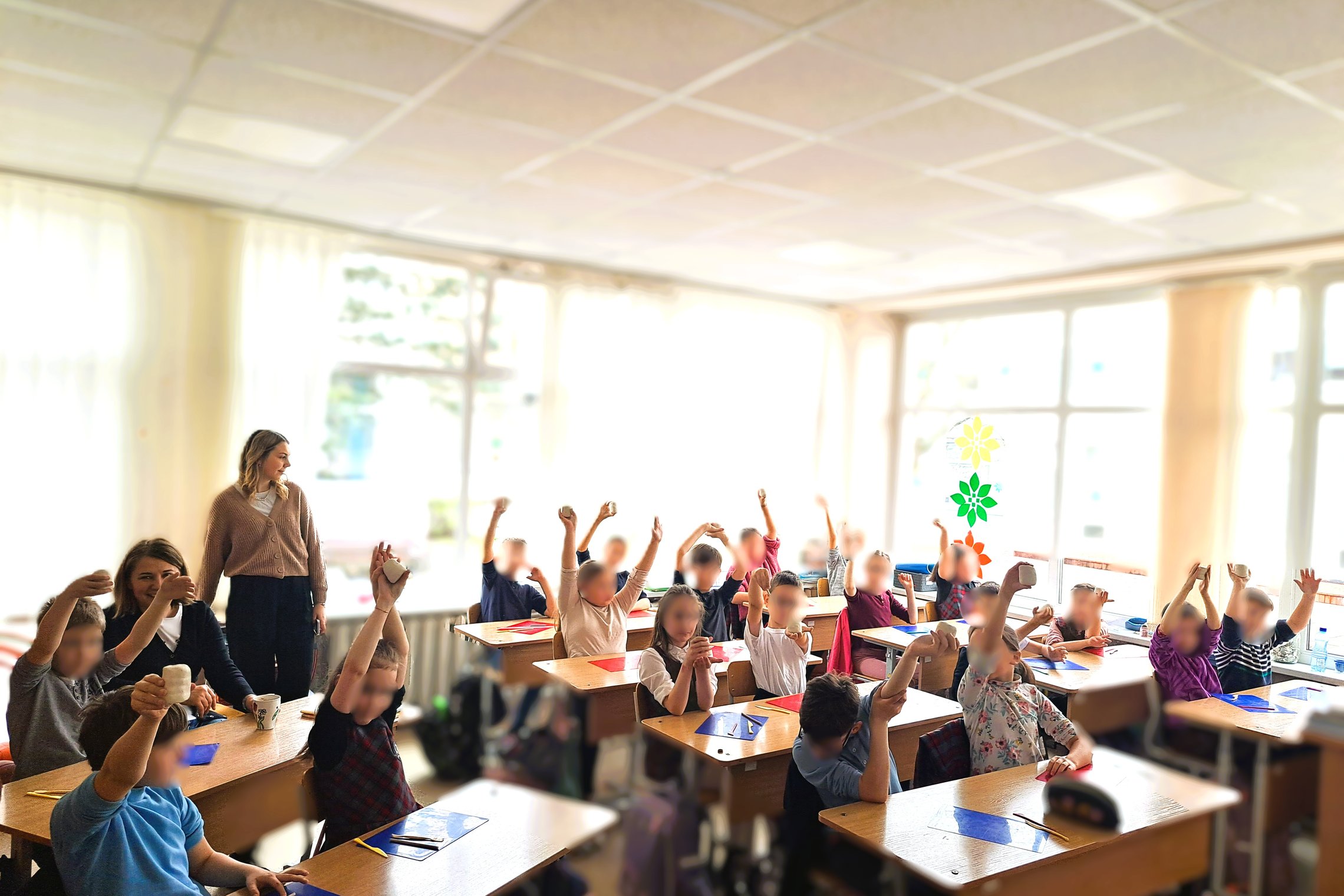 Šviesioje klasėje prie langų sėdi pradinukai, kurie laiko rankose iškeltas molio figūrėles. Mokytoja stebi vaikus. Ant langų matyti margaspalviai gėlių formos dekorai. Vaikai atrodo susikaupę ir džiaugiasi savo kūriniais.