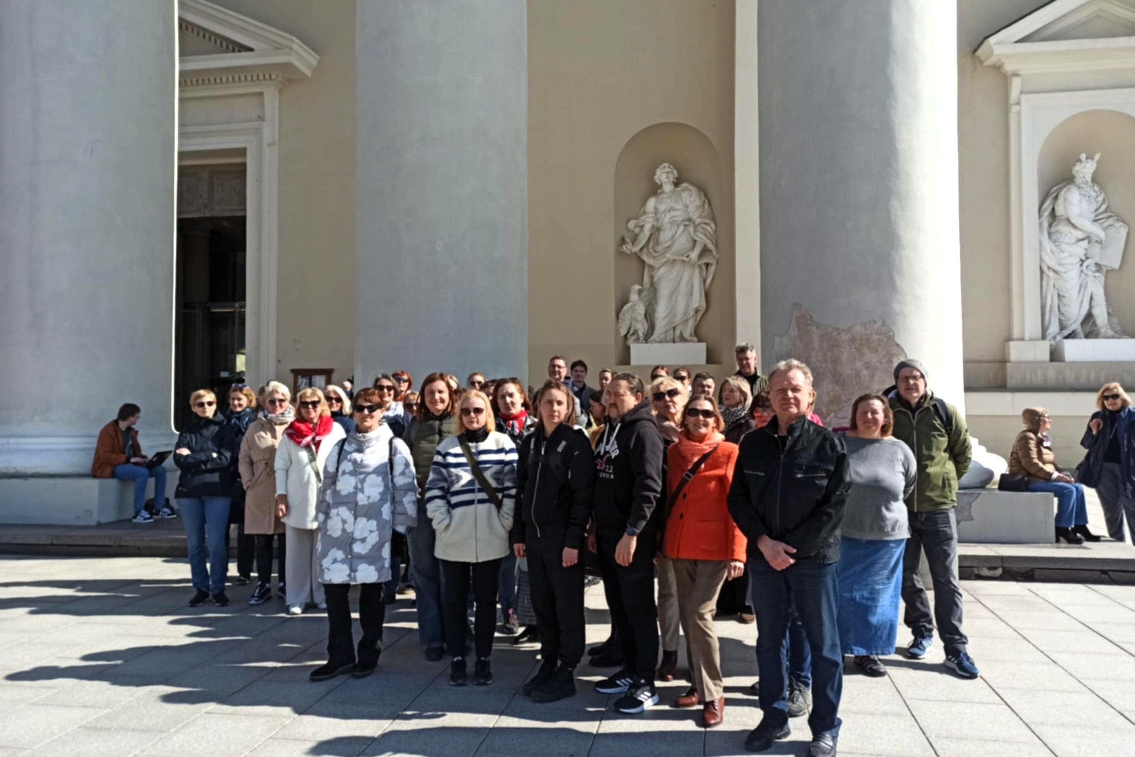Nuotraukoje užfiksuota didelė žmonių grupė, pozuojanti prie Vilniaus arkikatedros bazilikos. Fone matyti įspūdingi balti kolonos formos architektūriniai elementai bei religinės skulptūros nišose. Saulėtą dieną ekskursijos ar susitikimo dalyviai atrodo nus