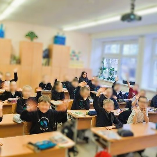 Pradinės mokyklos klasė su mokytoja ir mokiniais, sėdinčiais prie suolų, kai kurie mokiniai kelia rankas, klasėje šilta atmosfera, pro langus matyti žiemos vaizdas.