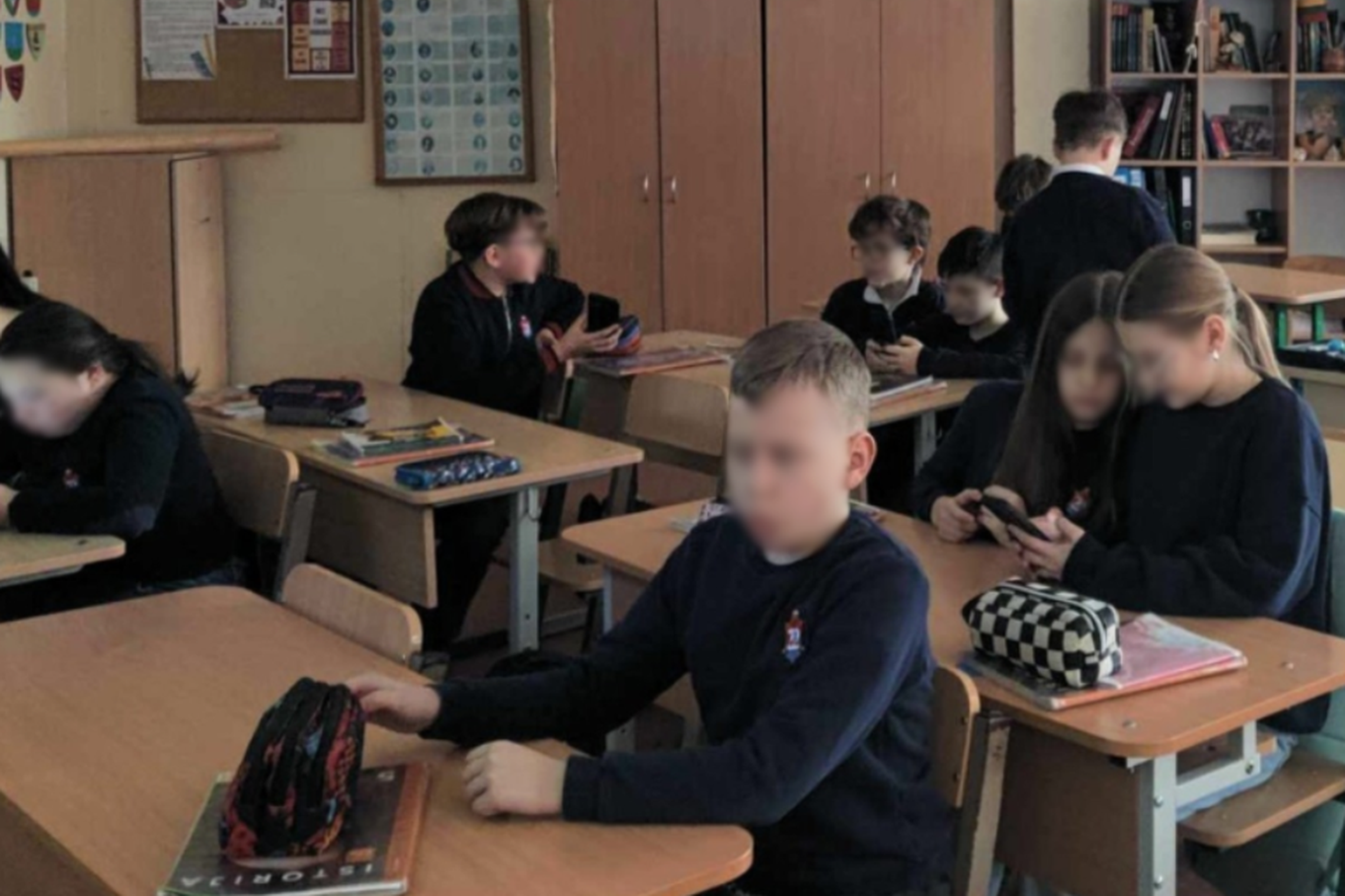 Šioje nuotraukoje matome mokyklos klasę, kurioje mokiniai sėdi grupelėmis arba po du prie stalų ir naudoja mobiliuosius telefonus. Dauguma jų dėvi tamsiai mėlynas ar juodas mokyklines uniformas su herbu ant krūtinės. Matomos lentynos su knygomis, kabinetų