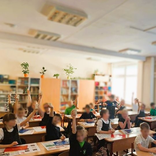 Klasėje prie suolų sėdintys mokiniai pakėlę rankas laiko molio gabalėlius, ant stalų ištiesti laikraščiai ir padėtos darbo priemonės, fone matyti lentynos su augalais ir mokymo priemonėmis, langai pridengti žaliuzėmis.