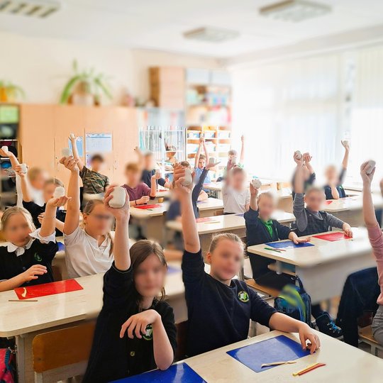 Klasėje prie suolų sėdintys mokiniai pakėlę rankas laiko baltos spalvos molio gabalėlius, ant stalų padėti spalvoti padėklai ir mediniai įrankiai, fone matyti lentynos su priemonėmis ir langai su vertikaliomis žaliuzėmis.