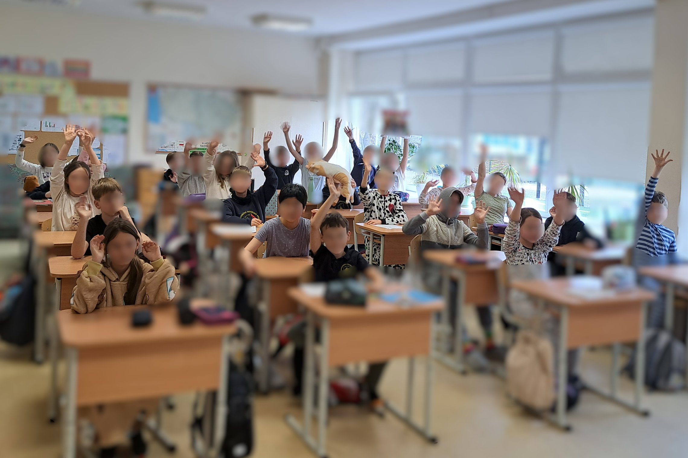 Klasėje prie savo suolų sėdi vaikai, dauguma pakėlę rankas ir mojuoja kamerai, vienas mokinys laiko pliušinį žaislą – šunį; šviesiame kambaryje matyti pasaulio žemėlapis ant sienos ir dideli langai su žaliuzėmis.
