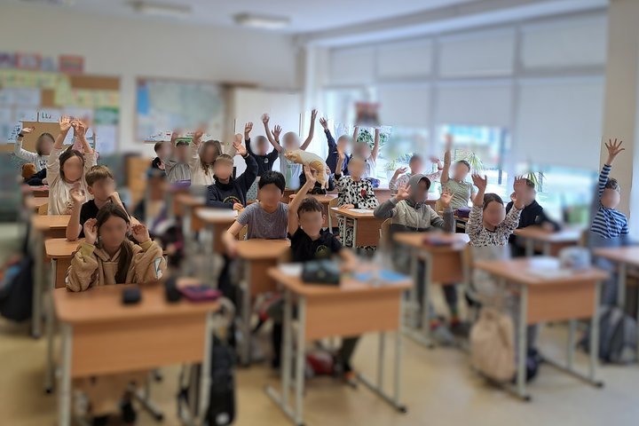 Klasėje prie savo suolų sėdi vaikai, dauguma pakėlę rankas ir mojuoja kamerai, vienas mokinys laiko pliušinį žaislą – šunį; šviesiame kambaryje matyti pasaulio žemėlapis ant sienos ir dideli langai su žaliuzėmis.