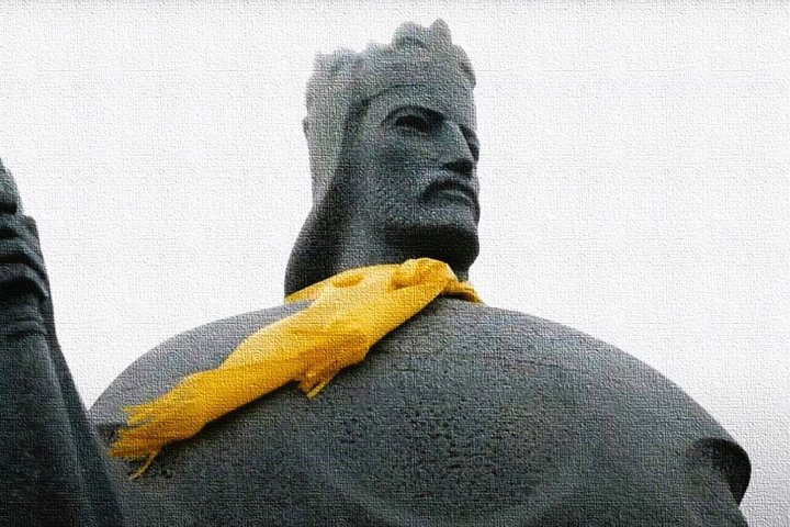 Paminklinė karaliaus Mindaugo figūra iš arti, ant kaklo užrištas geltonas šalikas, skulptūra perteikta tekstūros efektu, fonas pilkas ir neutralus.