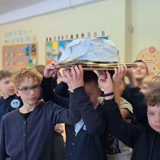 Grupė vaikų klasėje iškėlę virš galvų savo sukurtą maketą – kalvą ar piliakalnį, suformuotą iš kartono, popieriaus ir plėvelės; fone matyti edukacinės aplinkos detalės: spalvota informacinė lenta, durys ir plakatai su užrašais apie tvarius gyvenimo įgūdži