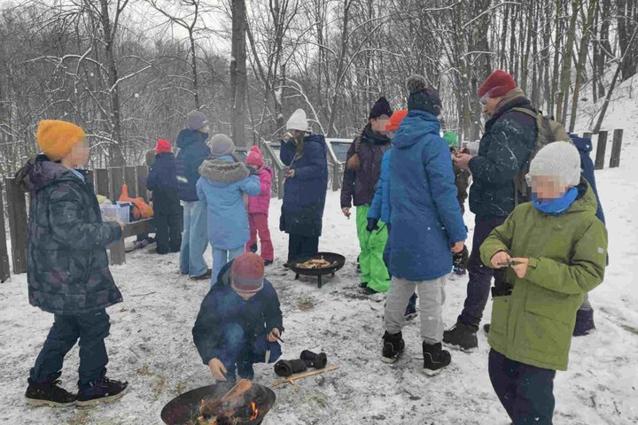 Nuotraukoje matoma grupė vaikų, dalyvaujančių lauko renginyje žiemos metu. Jie susirinkę aplink kelias ugnis, kuriose dega laužai. Kai kurie vaikai stovi ir šnekučiuojasi, kiti kepa užkandžius ant laužo. Aplinka apsnigta, o visi dėvi šiltus žiemos drabuži
