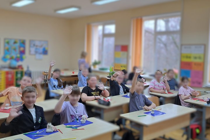 Pradinių klasių mokiniai sėdi klasėje prie stalų ir lipdo figūrėles iš molio. Ant stalų matyti plastilinui skirtos lentelės, įvairiaspalviai įrankiai ir jau suformuoti darbai. Daugelis vaikų pakėlę rankas mojuoja ar sveikinasi. Klasės fone matyti mokytoj