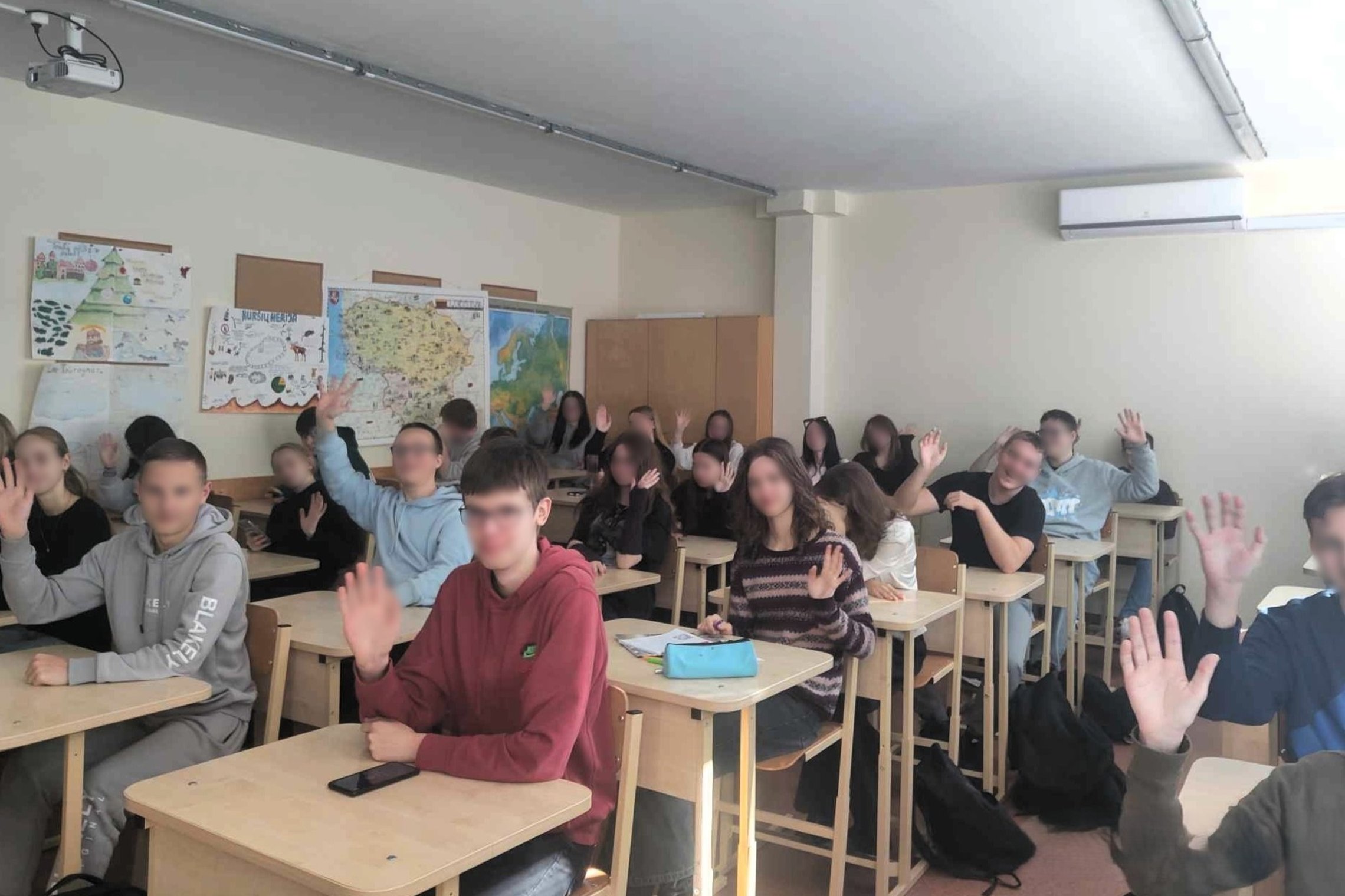 Vidurinės mokyklos mokiniai sėdi klasėje prie suolų ir mojuoja į kamerą, fone ant sienos kabo žemėlapiai ir mokinių darbai, o klasės interjeras pasižymi šviesiomis sienomis, mediniais baldais ir šiuolaikiška įranga.