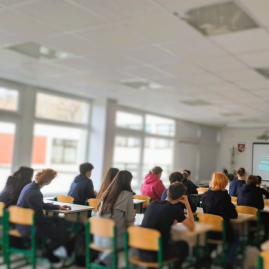 Mokiniai sėdi prie stalų ir žiūri į ekraną, rodomos skaidrės. Virš ekrano kabo Lietuvos herbas. Klasėje daug šviesos, nes pro langus sklinda natūrali dienos šviesa. Atmosfera rimta, mokiniai susikaupę.