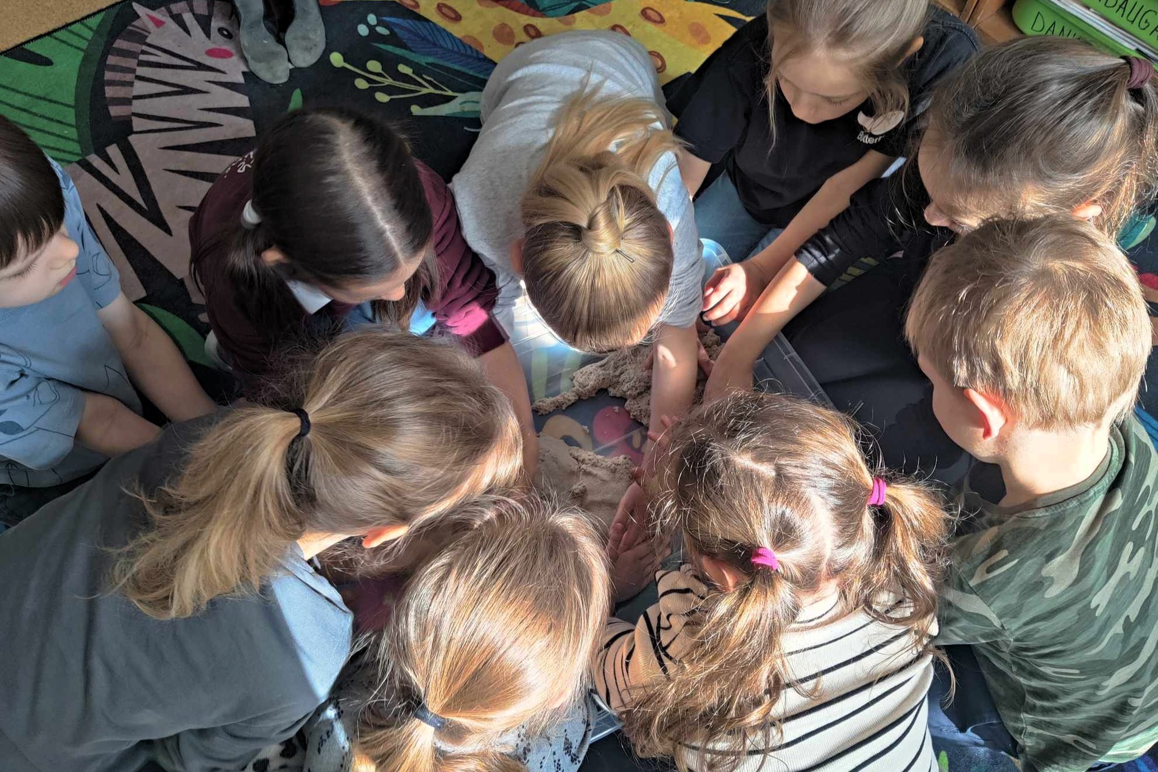 Grupė vaikų susibūrę ratu ir susikaupę stebi bei liečia kinetinį smėlį dėžėję, jie sutūpę ant spalvingo kilimo saulės nutviekstoje klasėje.