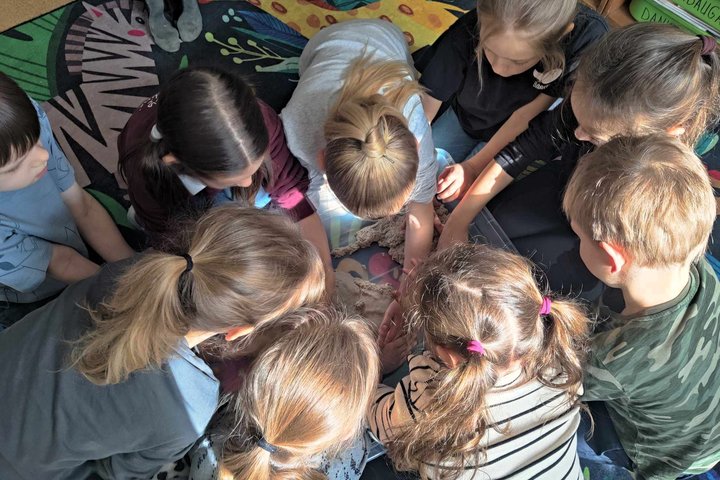 Grupė vaikų susibūrę ratu ir susikaupę stebi bei liečia kinetinį smėlį dėžėję, jie sutūpę ant spalvingo kilimo saulės nutviekstoje klasėje.