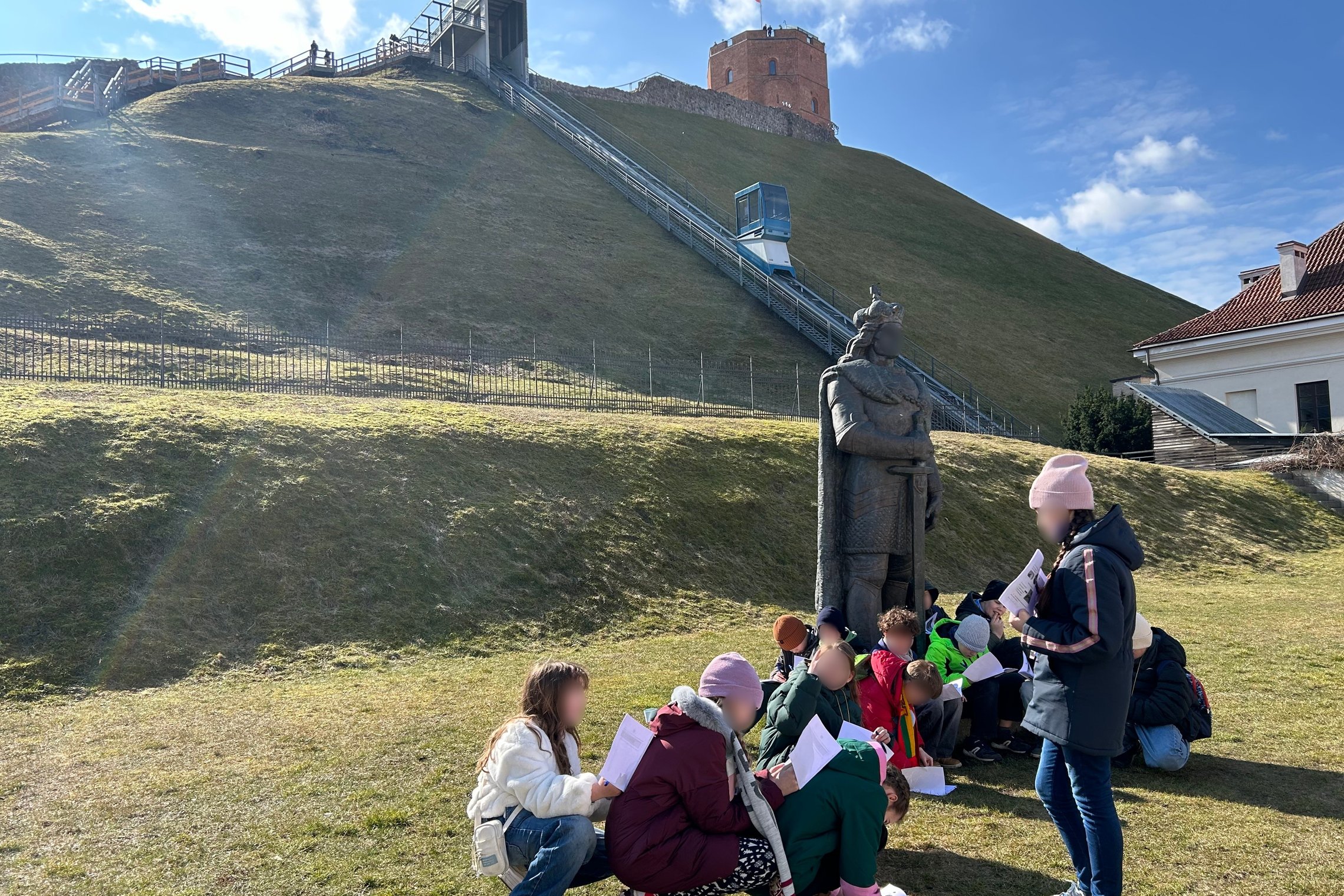 Nuotraukoje užfiksuota edukacinė veikla prie kunigaikščio Gedimino paminklo, esančio prie Vilniaus pilių kalno. Priekyje matoma grupė vaikų, susidomėję rašo ar skaito iš lapų, o viena mokytoja veda veiklą. Fone iškilęs Gedimino pilies bokštas su Lietuvos