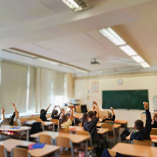 Mokiniai klasėje sėdi atsisukę į lentą ir pakėlę rankas su balsavimo kortelėmis ar daiktais, klasės viduje ryški šviesa, pro langus sklinda dienos šviesa, o priekyje matyti žalia lenta ir mokytojo stalas.
