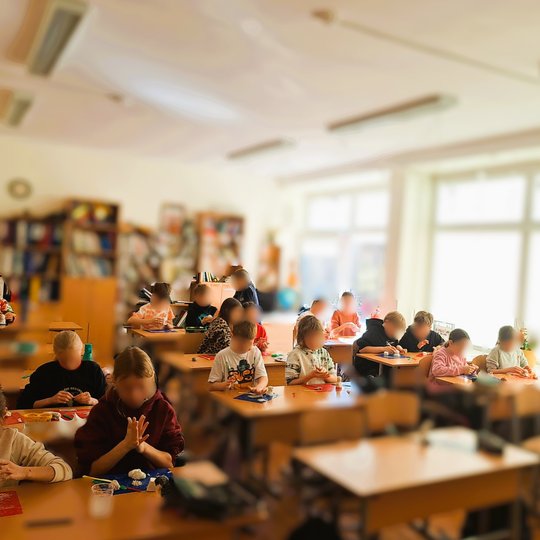 Šviesi pradinių klasių mokinių klasė, kurioje vaikai susikaupę lipdo figūras iš molio. Mokiniai sėdi prie stalų, ant kurių padėti raudoni, mėlyni ar kitos spalvos padėkliukai, o juose – įvairūs molio kūriniai. Už langų šviečia ryški dienos šviesa, kambary
